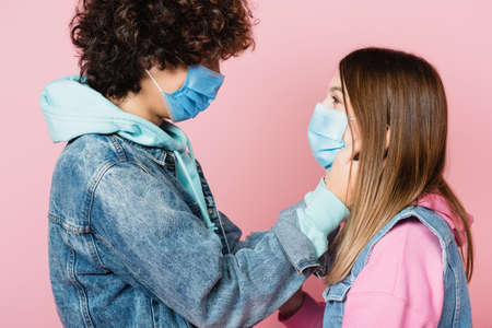 Teen couple in medical masks looking at each other isolated on pink