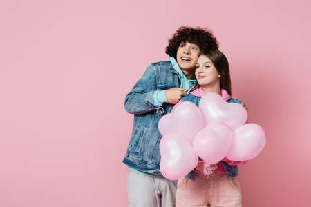 Cheerful teenager sanding near girlfriend with balloons isolated on pinkの写真素材