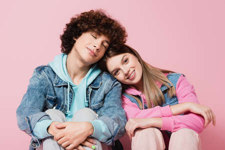 Smiling teenager looking at camera near boyfriend with closed eyes on pink background