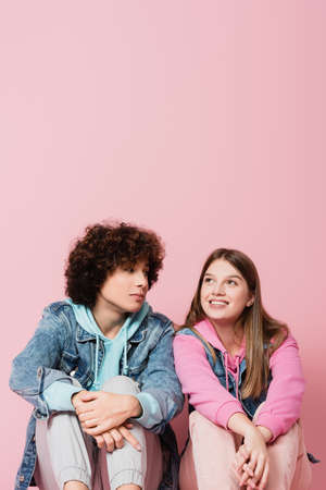 Curly teenager looking at smiling girlfriend on pink background