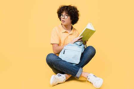 Curtly teenager with backpack holding book while sitting on yellow backgroundの写真素材