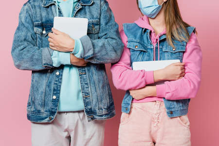 Cropped view of teenager in medical mask holding digital tablet near friend on pink background
