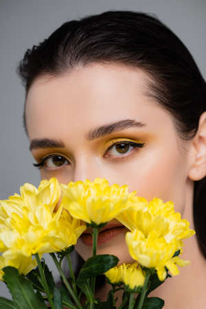 young woman with yellow eyeshadows covering face with blooming flowers isolated on grayの写真素材