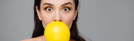young brunette woman with makeup blowing yellow bubblegum isolated on gray, banner