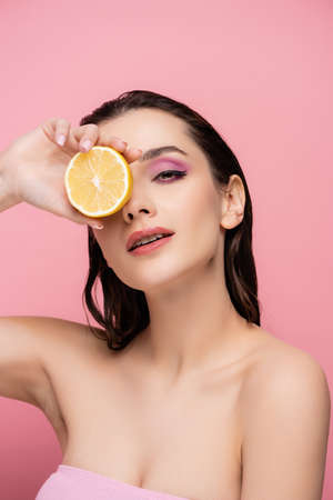young woman with bare shoulders covering eye with half of ripe lemon and looking at camera isolated on pinkの写真素材