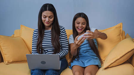 happy kid playing game on smartphone while mother using laptopの写真素材