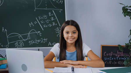 cheerful kid sitting at desk with laptop and looking at camera