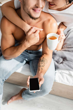 Cropped view of smiling woman with cup hugging boyfriend with smartphone on bedの写真素材