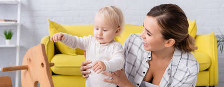 young cheerful woman supporting little boy standing near rocking horse, banner