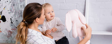 young woman showing toy bunny to child at home, banner