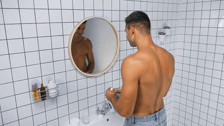 young shirtless man holding deodorant near mirror in bathroom