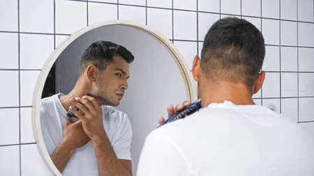 man in white t-shirt shaving neck with electric razor near mirror in bathroom