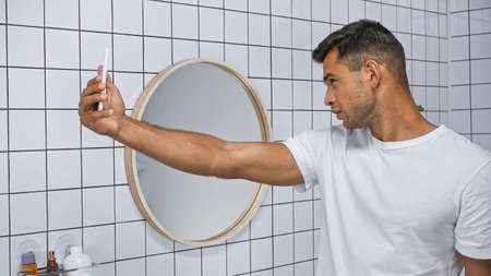 young man in white t-shirt taking selfie on mobile phone in bathroomの写真素材
