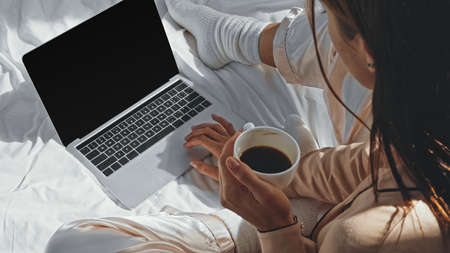 high angle view of woman holding cup of coffee near laptop with blank screen on bedの写真素材