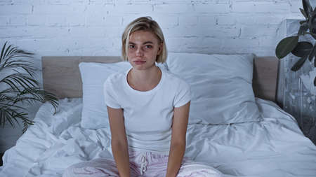 upset diseased woman in pajamas looking at camera while sitting in bedroom