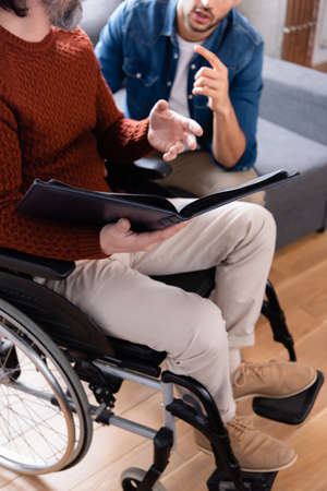 cropped view of young man pointing with finger near disabled father sitting with photo album in...