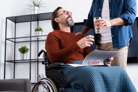 young man giving glass of water to smiling handicapped man sitting with newspaper in wheelchairの写真素材
