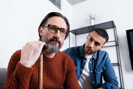 depressed man with walking stick near young hispanic son on blurred backgroundの写真素材