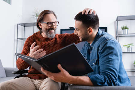 cheerful man touching head of happy hispanic son holding photo album on blurred foregroundの写真素材