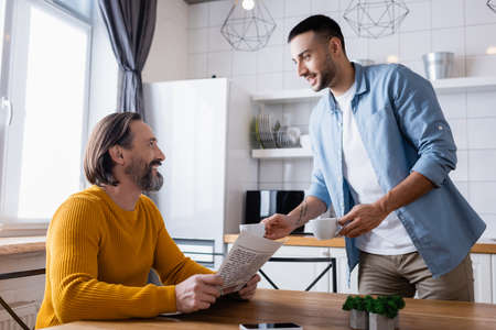smiling hispanic man holding coffee cups near father sitting with newspaper in kitchenの写真素材