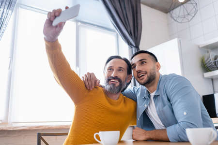 happy bearded man taking selfie with smiling hispanic son in kitchen