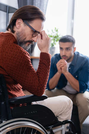 upset disabled man in wheelchair near young hispanic son on blurred backgroundの写真素材
