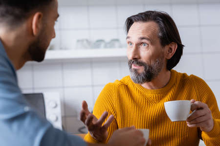 bearded man holding coffee cup and gesturing while talking to son in kitchen, blurred foregroundの写真素材