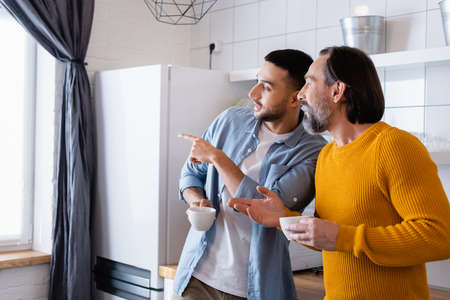 young hispanic man pointing with finger while looking away with father in kitchenの写真素材