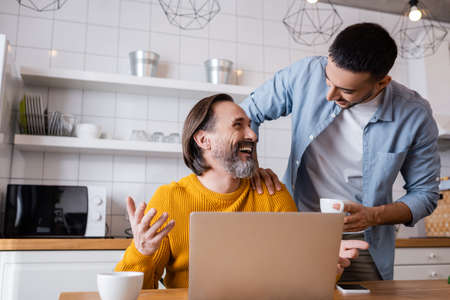 laughing man looking at young hispanic son while sitting near laptop in kitchen