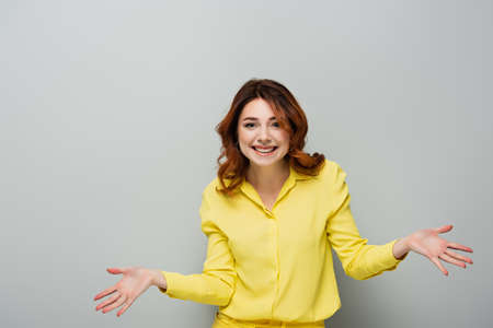 cheerful woman looking at camera while standing with open arms on grayの写真素材