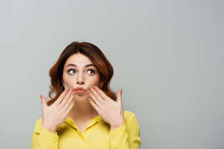 surprised woman looking away while touching cheeks isolated on grayの写真素材