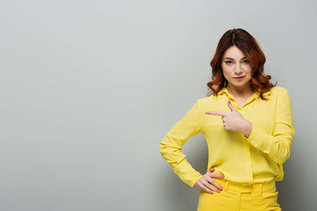 young curly woman in yellow shirt standing with hand on hip and pointing with finger on gray