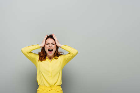 angry woman screaming while standing with closed eyes and touching head on gray