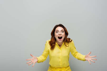 amazed woman looking at camera while standing with open arms on gray