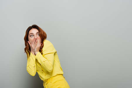 scared woman in yellow clothes covering mouth while looking away on gray