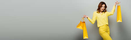 joyful woman looking at camera while holding yellow shopping bags on gray, bannerの写真素材
