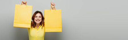 cheerful woman showing yellow shopping bags on gray, bannerの写真素材