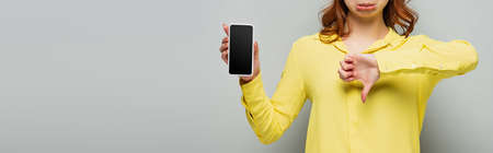 partial view of displeased woman showing dislike while holding smartphone with blank screen on gray, bannerの写真素材