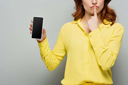 cropped view of woman showing hush gesture while holding mobile phone with blank screen on grayの写真素材