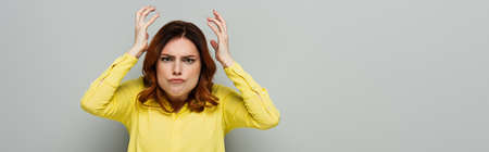 displeased woman looking at camera while holding hands near head on gray, bannerの写真素材
