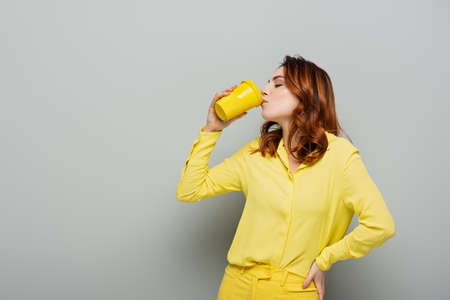 young woman standing with hand on hip and drinking coffee to go on gray