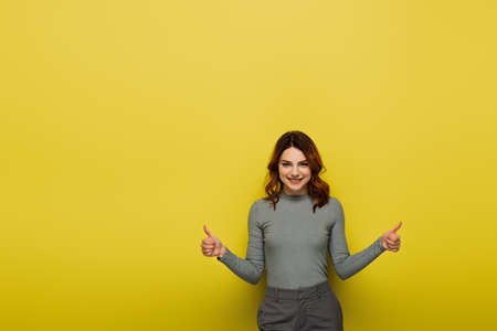 happy woman with curly hair showing thumbs up on yellowの写真素材
