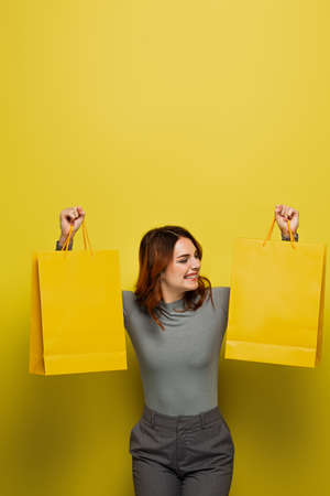 happy young woman with curly hair holding shopping bags on yellowの写真素材
