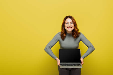 cheerful young woman with curly hair holding laptop with blank screen and biting lips on yellowの写真素材