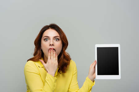 shocked woman covering mouth with hand while holding digital tablet with blank screen isolated on...