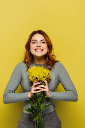 joyful young woman with wavy hair holding flowers on yellowの写真素材