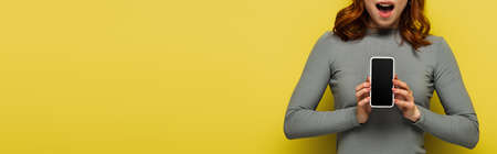 cropped view of amazed woman with holding smartphone with blank screen isolated on yellow, bannerの写真素材
