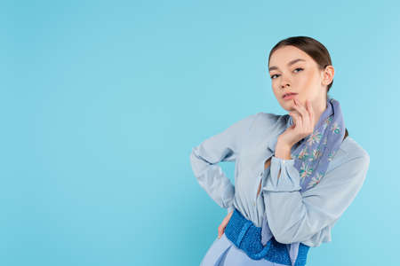 stylish woman touching face and looking at camera while standing with hand on hip isolated on blueの写真素材