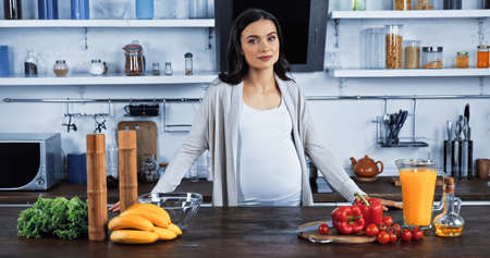 Pregnant woman looking at camera near fresh vegetables and orange juiceの写真素材