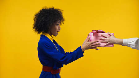 Side view of amazed african american woman taking gift box isolated on yellow
